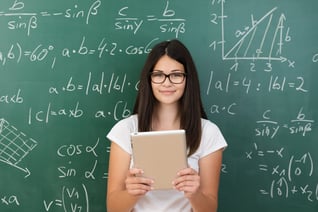 Clever attractive young female college student wearing glasses and holding a tablet computer in her hands standing in front of a chalkboard covered in equations in maths class smiling at the camera